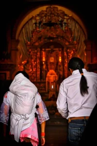 Back view of an indigenous man and woman in traditional dress walking toward a glowing, ornate gold church altar for the baptism of their child