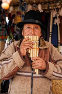 A portrait of an Ecuadorian man in a black fedora playing a wooden zampoña pan flute at a market stall.