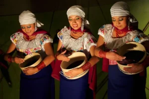 Three Ecuadorian women in traditional white embroidered blouses and blue skirts performing a cultural dance with wooden sieves.