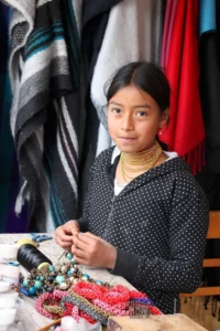 A young Ecuadorian girl with gold beaded necklaces sitting at a table making handmade jewelry in a workshop