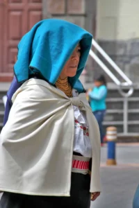Profile portrait of an older Ecuadorian woman wearing a blue headcover, cream shawl, and traditional gold beaded necklaces.