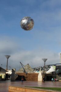The spherical silver "Ferns" sculpture suspended in the air over the stone steps and modern buildings of Wellington's Civic Square.