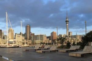 Auckland city skyline at dusk with the Sky Tower, harbor sailboats, and wooden lounge chairs on a pier.