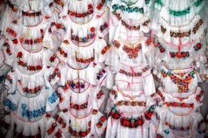 Traditional white Kichwa blouses with intricate, colorful floral embroidery displayed vertically in Otavalo