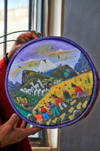 A local artist holding a circular sheepskin painting depicting a colorful highland scene with a volcano and llamas.